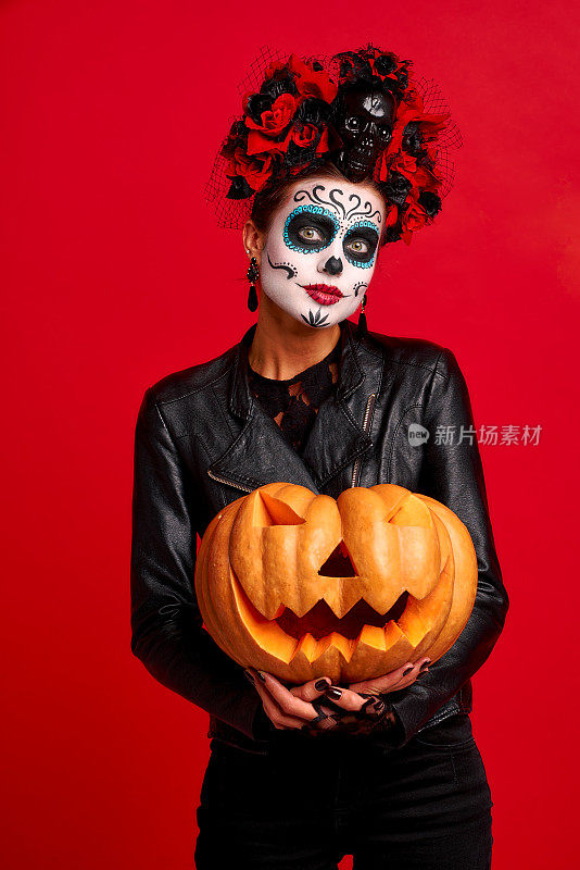 万圣节派对女孩。Happy girl with sugar skull makeup, with a花环of flowers on her head and skull, wearing lace gloves and leather jacket . Happy girl with sugar skull makeup, with a花环of flowers on her head and skull, wearing lace gloves and leather jacket . Happy girl with sugar skull makeup, with a花环of flowers on her head and skull, wearing蕾丝手套and leather jacket, Holds a big Jack-o-lantern pumpkin isolated on red